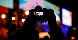 A person holds up a camera at night, capturing a brightly lit scene with colorful lights and blurred shapes in the background. The camera’s display screen shows the focused image being taken.