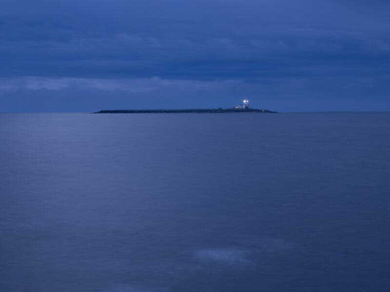 A distant island with a lighthouse glowing faintly, surrounded by calm, blue water under a cloudy twilight sky.
