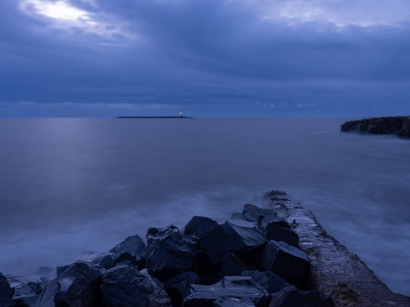 Rocky coastline at dusk with calm, misty water in the foreground and a distant island featuring a small lighthouse. The sky is filled with dramatic, swirling clouds in blue tones.