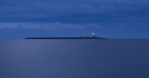 Coquet island with a lighthouse illuminated at dusk, surrounded by calm, blue ocean water under a cloudy sky.