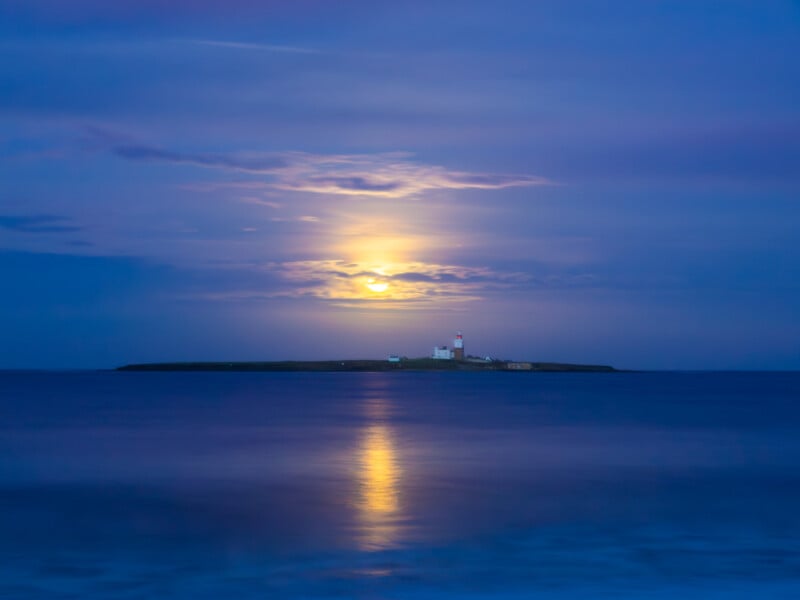 A small island with a lighthouse sits in calm blue water under a cloudy sky, with a bright full moon shining and reflecting on the sea.