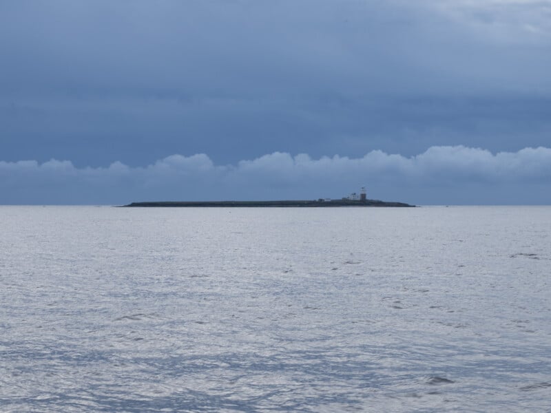 Coquet island with its lighthouse stands in the distance, surrounded by calm, silvery-blue sea under a cloudy, overcast sky.