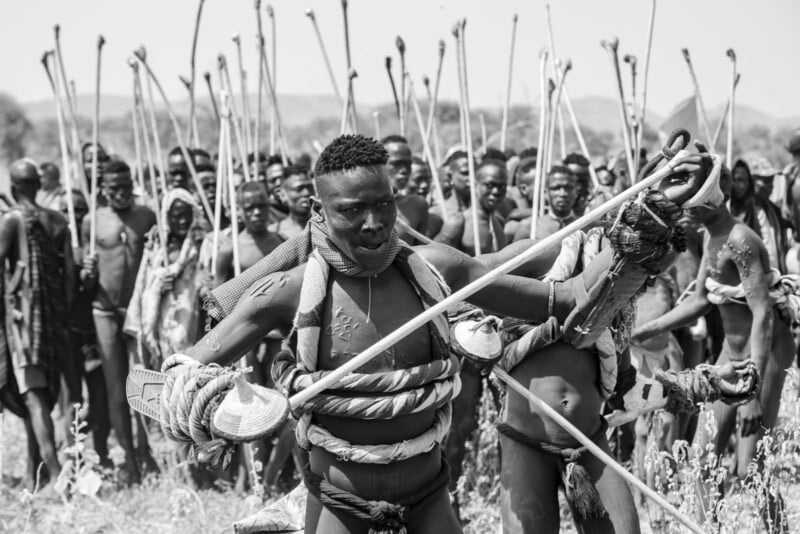 A group of men, some with body paint and decorative ropes, stand outdoors holding long sticks or staffs, with one man in front performing a dynamic gesture. The scene appears ceremonial or traditional.