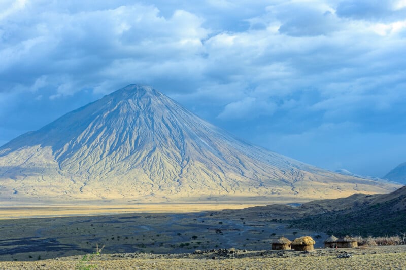 A towering, conical mountain rises above a vast, dry plain under a cloudy sky, with several small thatched huts clustered in the foreground.