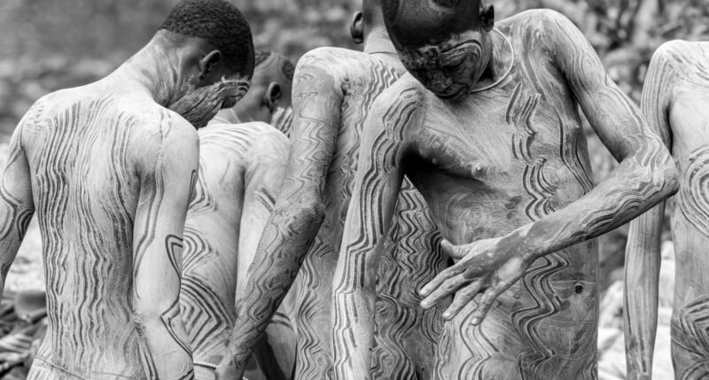 Black and white photo of several people with intricate body paint designs covering their skin, standing closely together with their backs and sides visible.
