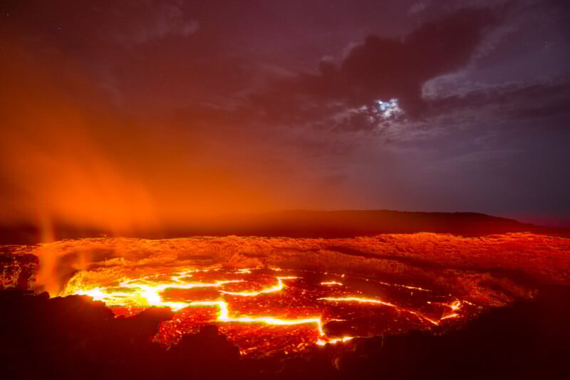 Molten lava glows bright orange and red inside an active volcanic crater at night, with steam rising and clouds partially obscuring the moon in the dark, dramatic sky.