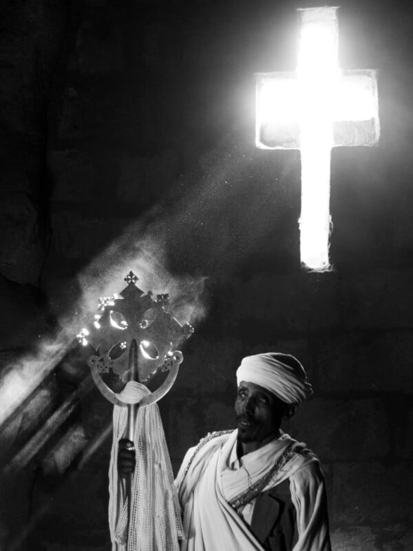 A man in traditional Ethiopian clothing holds an ornate cross, standing in front of a stone wall with sunlight streaming through a glowing cross-shaped window above him. The image is in black and white.