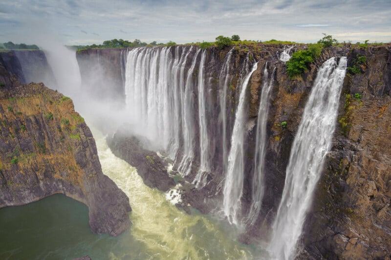 A wide, dramatic waterfall cascades over a rocky cliff surrounded by lush green vegetation under a cloudy sky, creating mist at the base where the water crashes into a river.