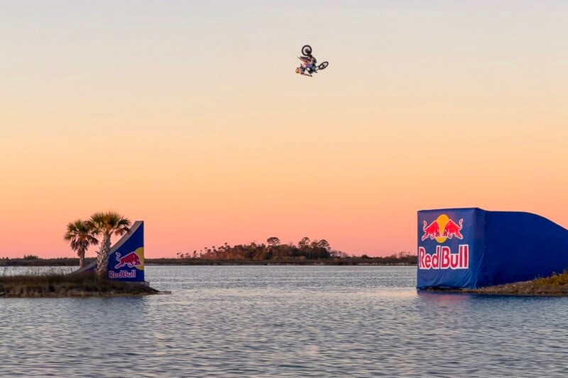 A motorcyclist performs a high jump over water at sunset, launching from a Red Bull-branded ramp on the right and aiming for a landing ramp across the water, with palm trees and an island in the background.