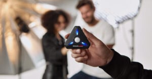 A person holds a black, triangular device with blue lights, focusing on it. Behind, two people are blurred, with professional lighting equipment in the backdrop. The device is central to the image.