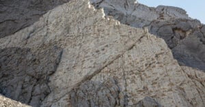 Large rocky cliff face with visible diagonal cracks and numerous small, round indentations covering some of the lighter rock surface; the background shows more rugged gray stone.
