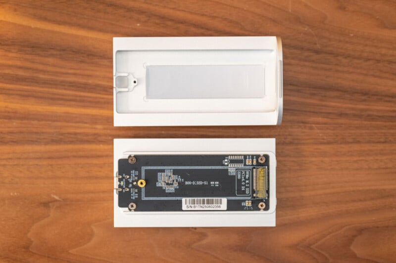 A white rectangular electronic device is shown open on a wooden surface. The top cover is removed, revealing a circuit board with electronic components and a barcode inside the lower part of the device.