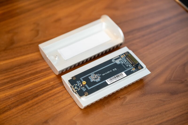 A solid-state drive (SSD) enclosure is open on a wooden surface, showing the internal circuit board and components, with the enclosure cover placed beside it.
