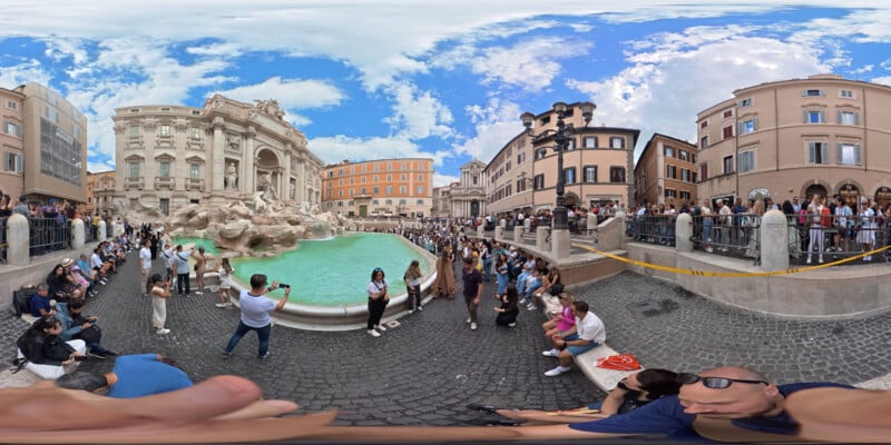 A wide, 360-degree view of the crowded Trevi Fountain in Rome shows tourists taking photos, tossing coins into the turquoise water, and admiring the historic baroque architecture under a partly cloudy blue sky.