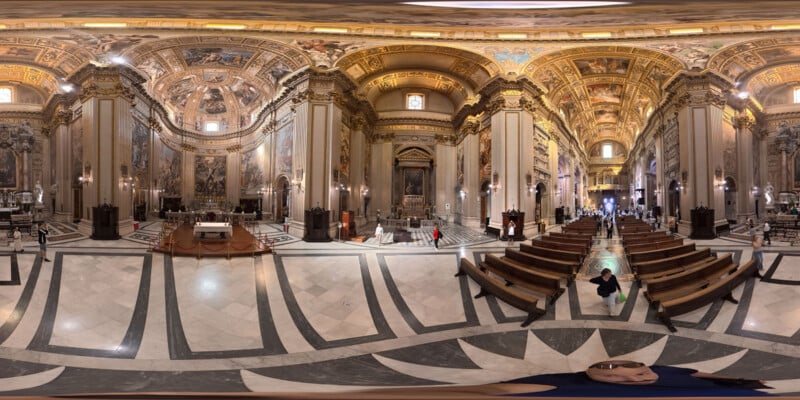 A panoramic view of an ornate cathedral interior with high vaulted ceilings, detailed frescoes, marble columns, and rows of wooden pews. Visitors walk and sit inside, admiring the grand architecture and decorative artwork.