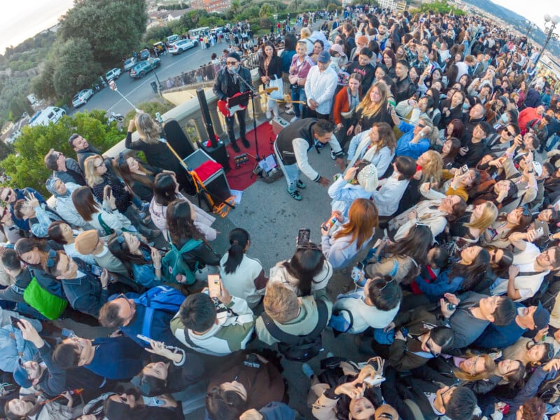 A large crowd forms a wide circle around street musicians performing outdoors; many people are taking photos and videos with their phones, capturing the lively scene on a sunny day.
