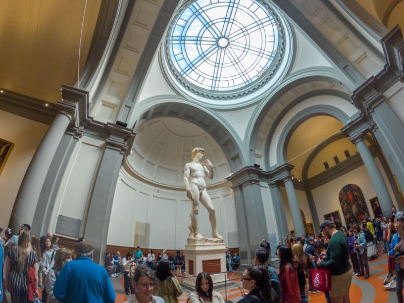 Visitors gather around Michelangelo’s statue of David, displayed under a large dome with a circular skylight in a spacious museum gallery. The room is filled with natural light and architectural details.