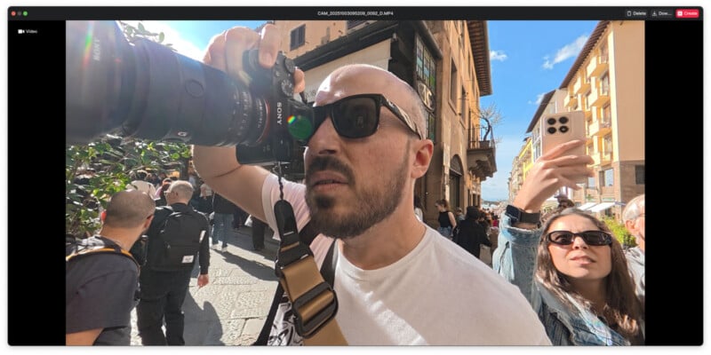 A man wearing sunglasses takes a photo with a professional camera on a sunny city street, while a woman behind him holds up a smartphone, also taking pictures. Other people walk in the background.