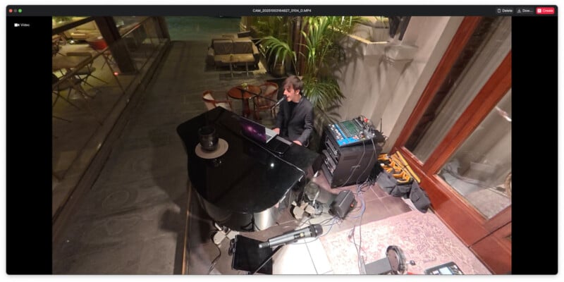 A person in a black suit plays a grand piano indoors near a mixing board and musical equipment, with tables, chairs, and potted plants visible through nearby windows.
