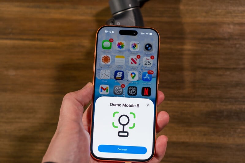 A hand holds an iPhone displaying a prompt to connect to "Osmo Mobile 6," with various app icons visible on the screen; a gimbal device is seen on a wooden surface in the background.