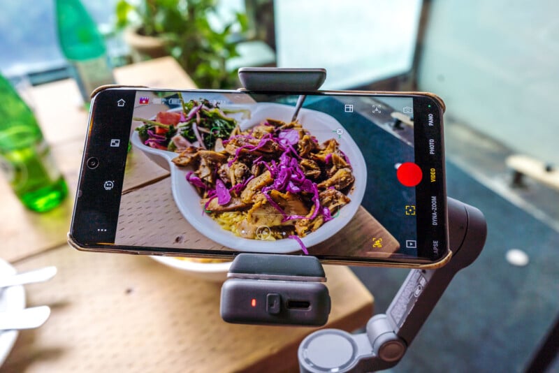 A smartphone on a stabilizer films a bowl of food with grilled meat, purple cabbage, and greens on a wooden table, with utensils and a green bottle nearby.
