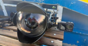A large underwater camera housing with a dome port and attached small action camera rests on a wooden bench, with a blue and yellow boat structure in the background. A person is reflected in the dome port.