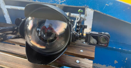 A large underwater camera housing with a dome port and attached small action camera rests on a wooden bench, with a blue and yellow boat structure in the background. A person is reflected in the dome port.
