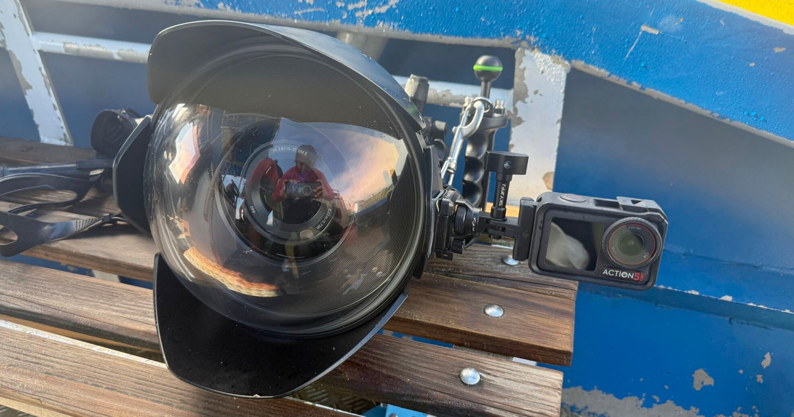 A large underwater camera housing with a dome port and attached small action camera rests on a wooden bench, with a blue and yellow boat structure in the background. A person is reflected in the dome port.