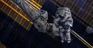 An astronaut in a spacesuit works outside the International Space Station, surrounded by large solar panels and equipment, with the darkness of space in the background.