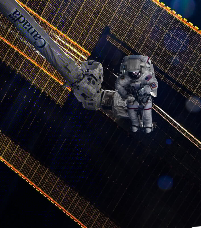 An astronaut in a spacesuit works on the end of the Canadarm2 robotic arm outside the International Space Station, with solar panels and the darkness of space in the background.
