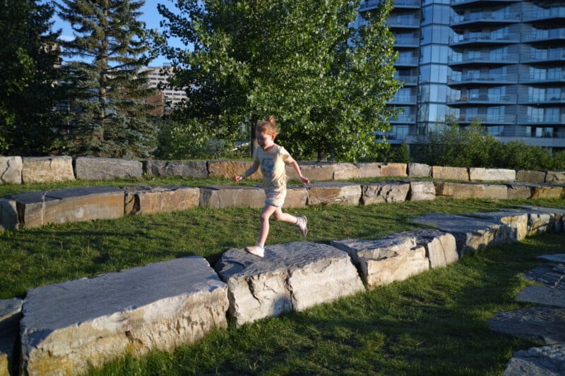 A young child runs along large stone slabs in a park, with grass, trees, and modern apartment buildings in the background on a sunny day.