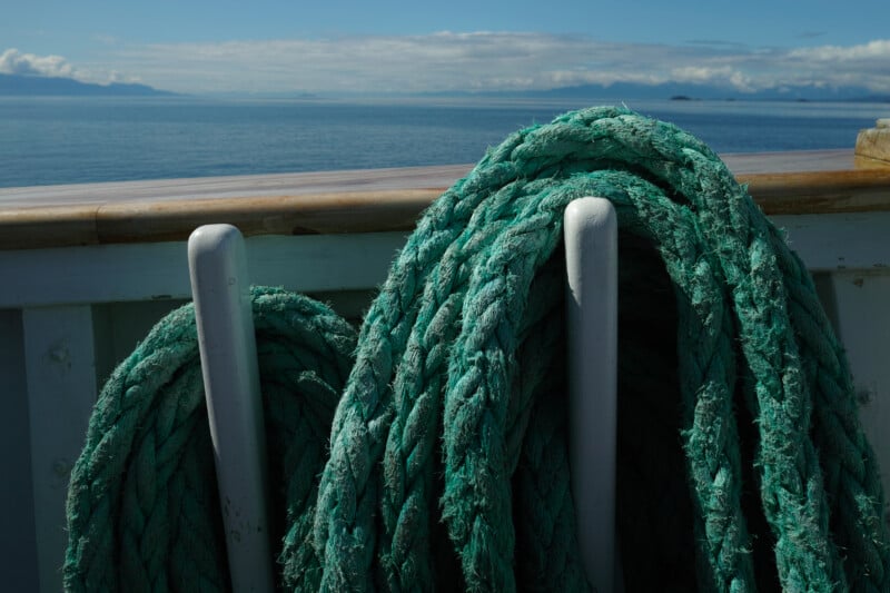 Thick green ropes coiled around white posts on a boat deck, with calm blue water and distant mountains visible under a cloudy sky in the background.