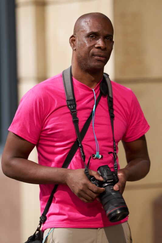 A person wearing a bright pink t-shirt holds a camera with both hands. They have camera straps around their neck and blue headphones hanging from their collar. The background is blurred and neutral-toned.