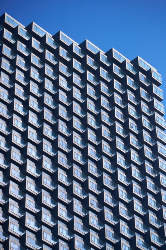 A modern high-rise building with repeating rows of balconies and windows, reflecting the blue sky, creating a geometric pattern against the clear backdrop.