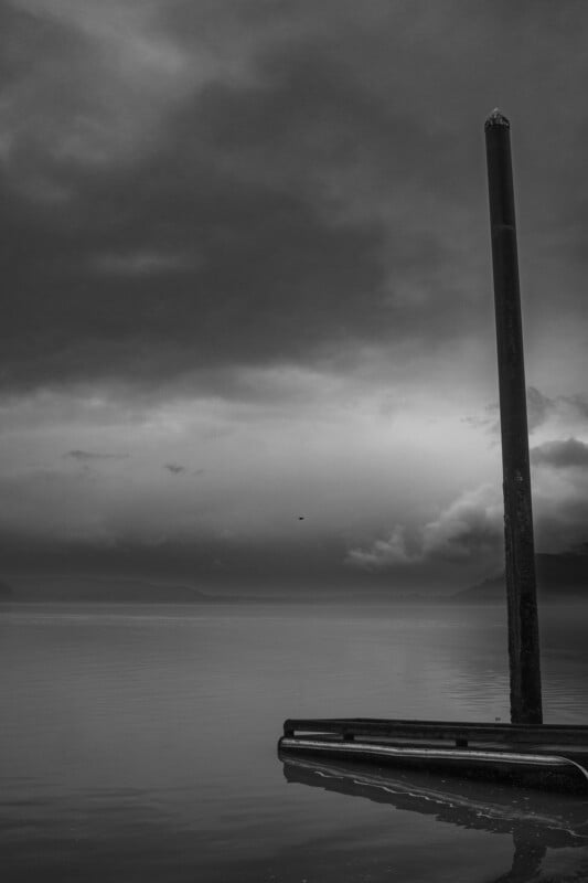 A tall pole rises from a calm, reflective body of water, next to a small dock. Dark clouds fill the moody sky, and distant mountains are barely visible through the mist. The scene is in black and white.