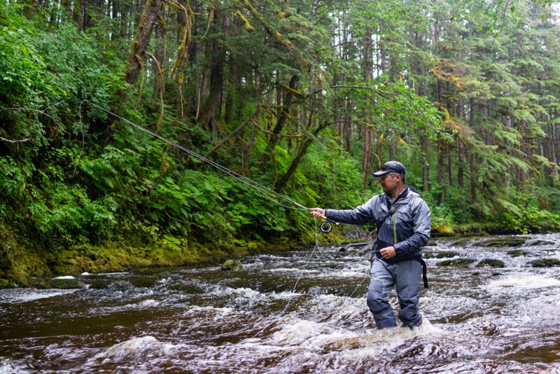 A man wearing waterproof gear stands in a flowing river surrounded by dense, green forest, fly fishing with a rod and line amid the rushing water.