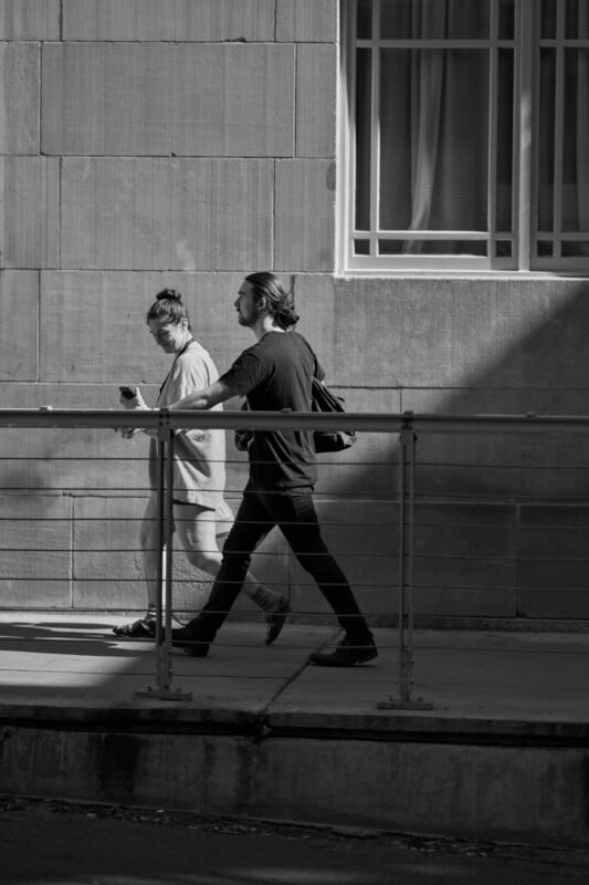 Two people walk side by side on a sidewalk next to a building with large windows. One holds a cup, and both are dressed casually. The scene is in black and white.