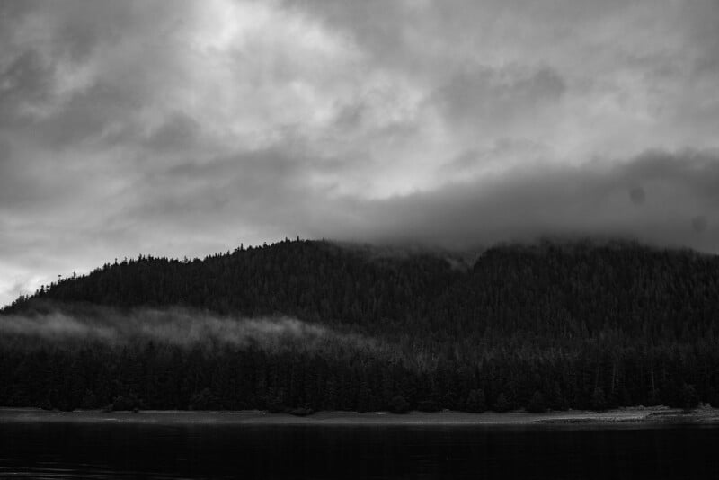 A dense forested hillside is shrouded in fog and low clouds under a moody, overcast sky. The scene is reflected on a calm body of water in the foreground. The photo is in black and white.
