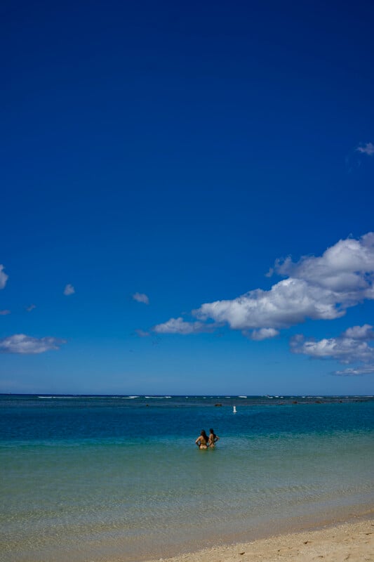 Two people stand in the shallow turquoise water of a beach under a clear, deep blue sky with a few scattered clouds. Sandy shore is visible in the foreground.