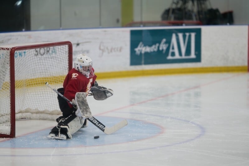 A youth hockey goalie in red gear is crouched in front of the net, focused on stopping a puck sliding on the ice toward the goal during a game at an indoor rink.