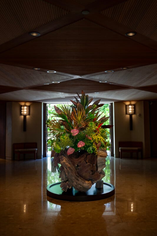 A large, vibrant floral arrangement with tropical flowers and greenery sits on a round pedestal in the center of a dimly lit, elegant lobby with wood accents and a marble floor. Light streams in from a window behind it.