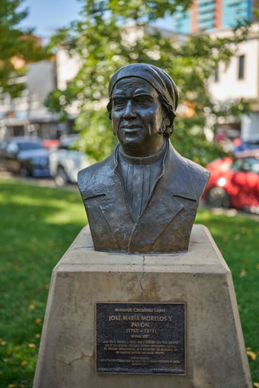 A bronze bust of José María Morelos y Pavón on a stone pedestal with a plaque, set outdoors on a grassy area, with trees, parked cars, and buildings visible in the background.