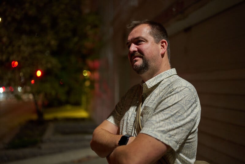 A man with short hair and a beard stands outdoors at night with his arms crossed, wearing a light patterned shirt. Blurred streetlights and trees appear in the background.