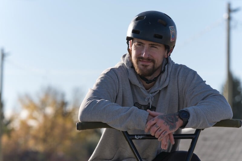 A man wearing a black helmet and gray hoodie sits on a BMX bike, smiling slightly. He has tattoos on his hands and a watch on his wrist. The background shows blurred trees and power lines.