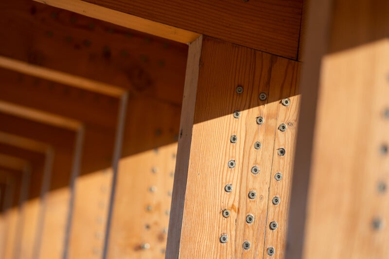 Close-up of a wooden structure with metal screws arranged in rows, showing strong light and shadow patterns across the smooth, warm-toned wood surfaces.