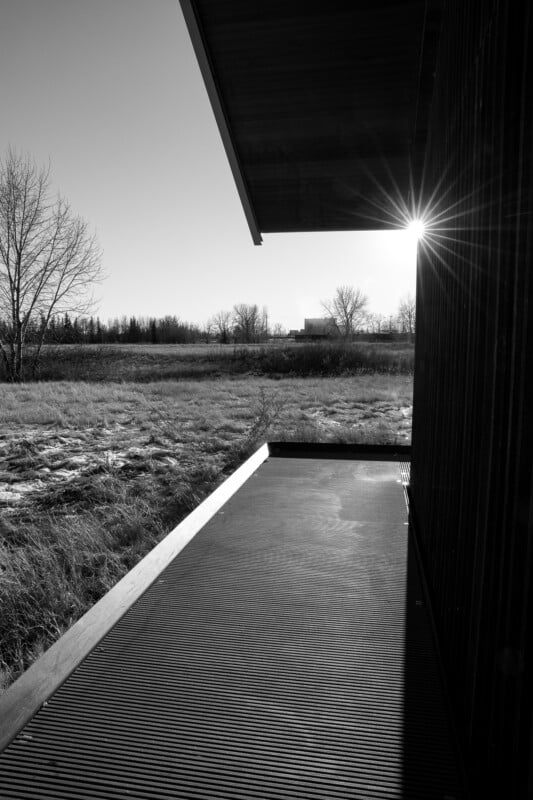 A black and white photo of a wooden deck next to a modern building, with leafless trees and grass in the background. The sun is low in the sky, creating a visible sunburst near the roof’s edge.