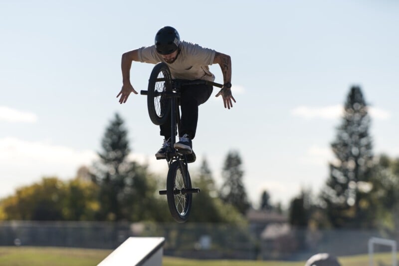 A person wearing a helmet performs a trick on a BMX bike in mid-air above a ramp at an outdoor skatepark, with trees and a fence visible in the background.