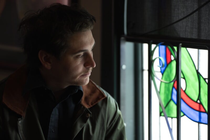 A man with dark hair and a dark jacket sits indoors, looking thoughtfully out of a window featuring colorful stained glass with green and blue patterns. Soft light illuminates his face.