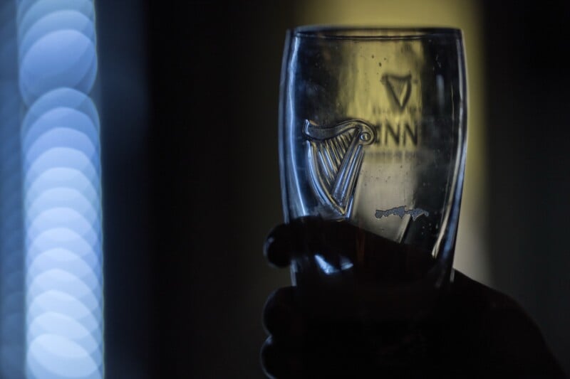 A hand holds a nearly empty Guinness glass in dim lighting, with blue and yellow blurred lights in the background. The glass features the Guinness harp logo and some text.