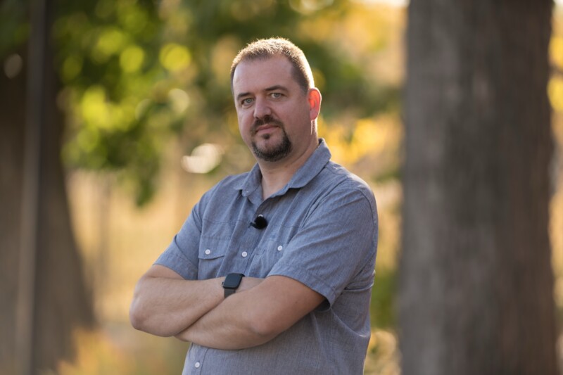 A man with short hair and a beard stands outdoors with arms crossed, wearing a gray short-sleeve button-up shirt and a smartwatch. The background is blurred with trees and sunlight.